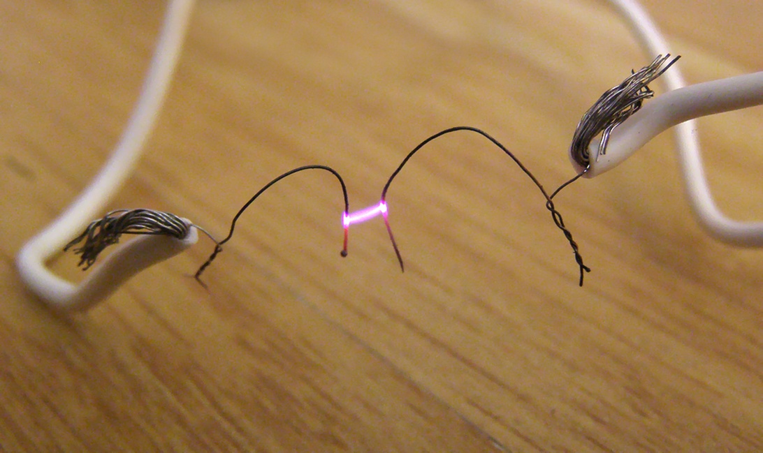 Närbild på skadade elkablar med fransiga ändar som visar en rosa/lila elektrisk ljusbåge mellan dem på en träyta.
